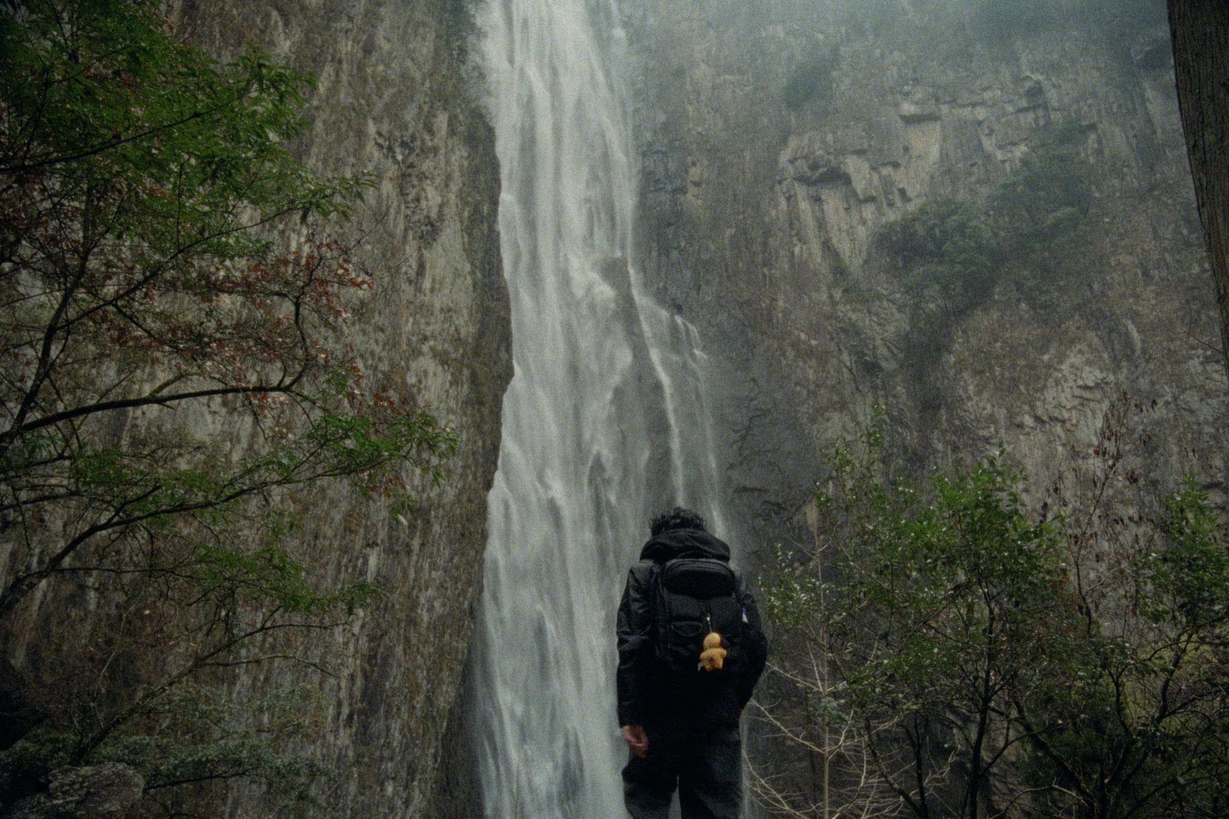 Andrew Amisola in People Who Meet Will Meet Again — Andrew from behind with a backpack and yellow duck keychain, facing a towering waterfall streaming down a sheer cliff face framed by trees.