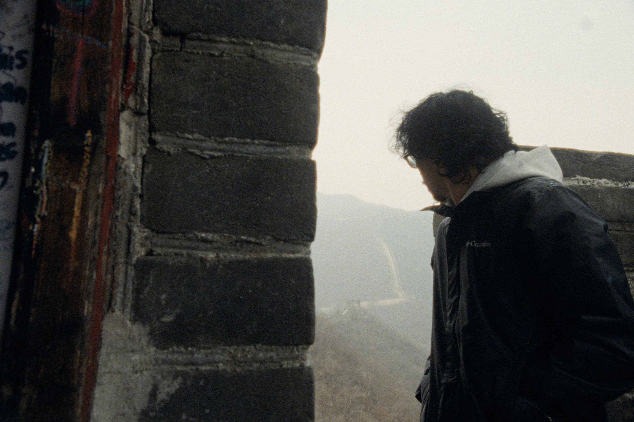 Andrew Amisola in People Who Meet Will Meet Again — Andrew looking down through an opening in the Great Wall, the Wall descending into hazy mountains behind him, colorful graffiti on the brick edge.