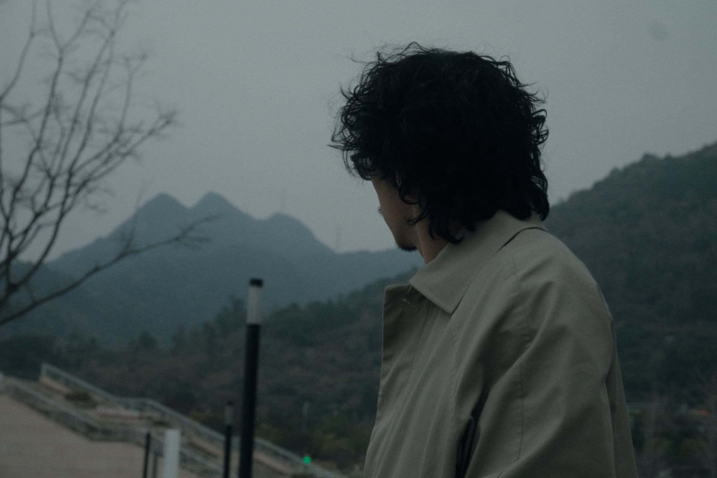 Andrew Amisola in People Who Meet Will Meet Again — Andrew from behind in a beige trench coat, curly hair wind-blown, looking down with misty mountains rising in the background.