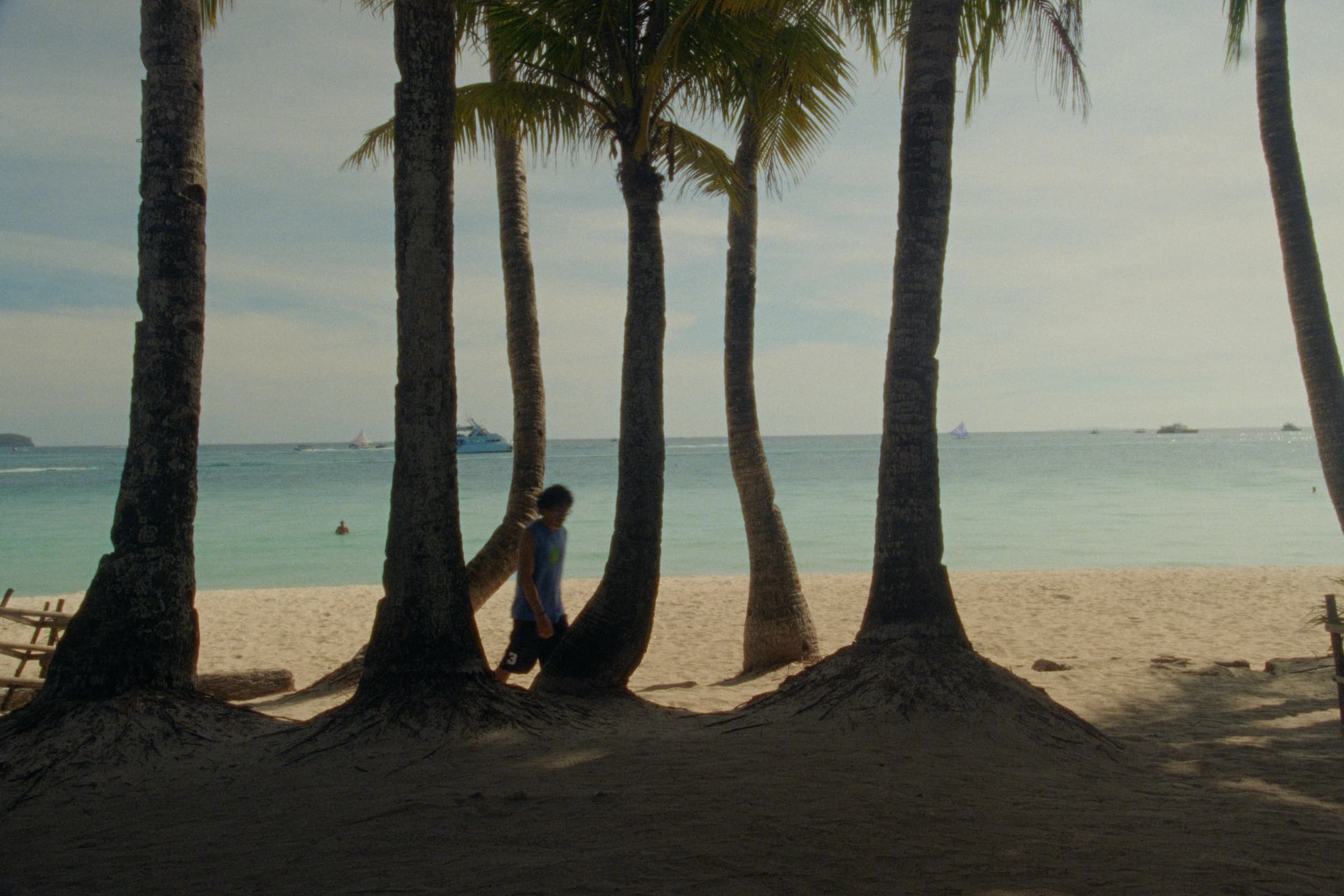 Andrew Amisola in White Sands in Boracay — A row of palm trunks rises from the sand with Andrew leaning against one of them, boats and bathers scattered across the turquoise sea behind.