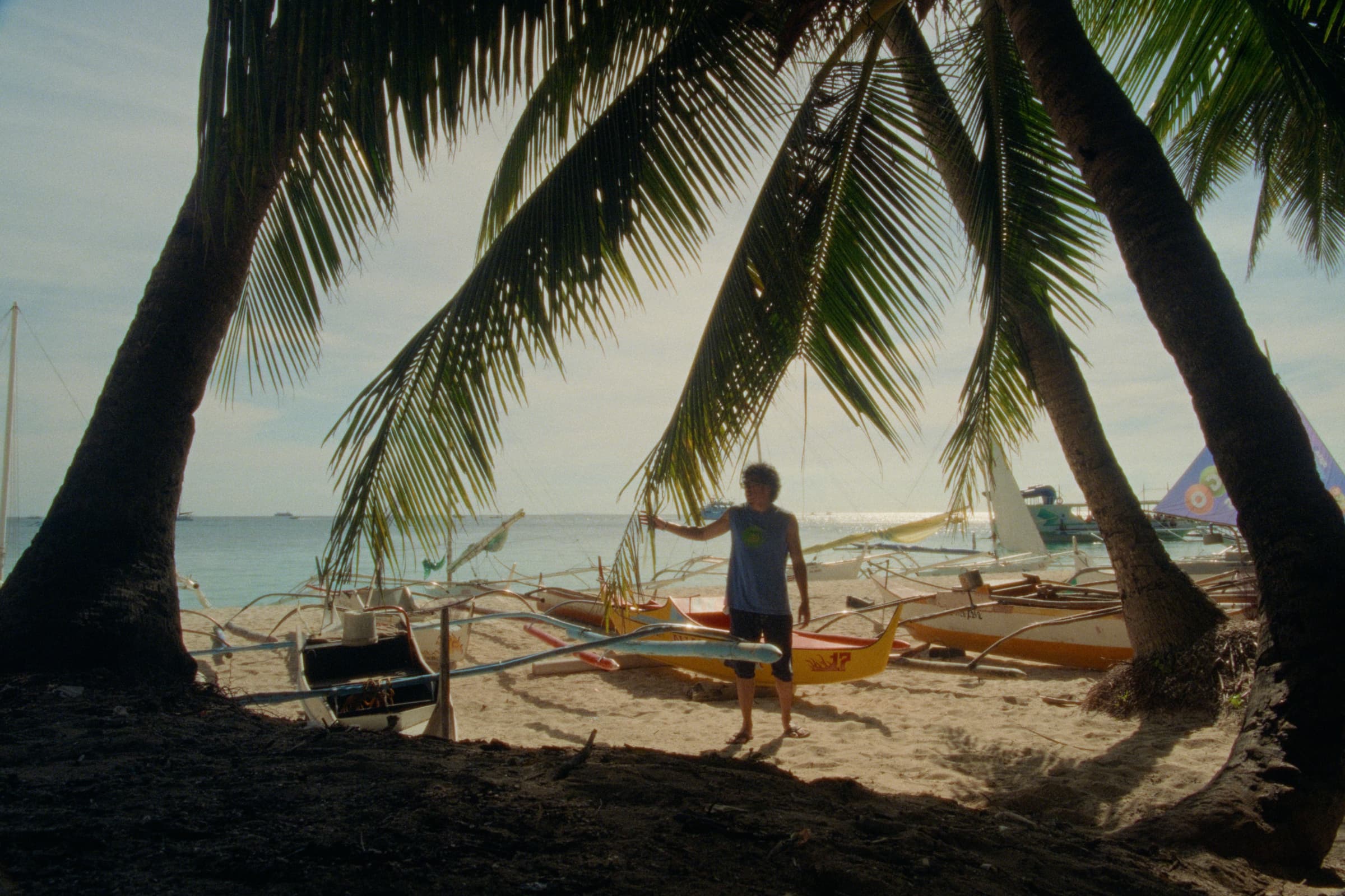 Andrew Amisola in White Sands in Boracay — Andrew standing beside a row of paraws on the sand, palm fronds dipping into the top of the frame.
