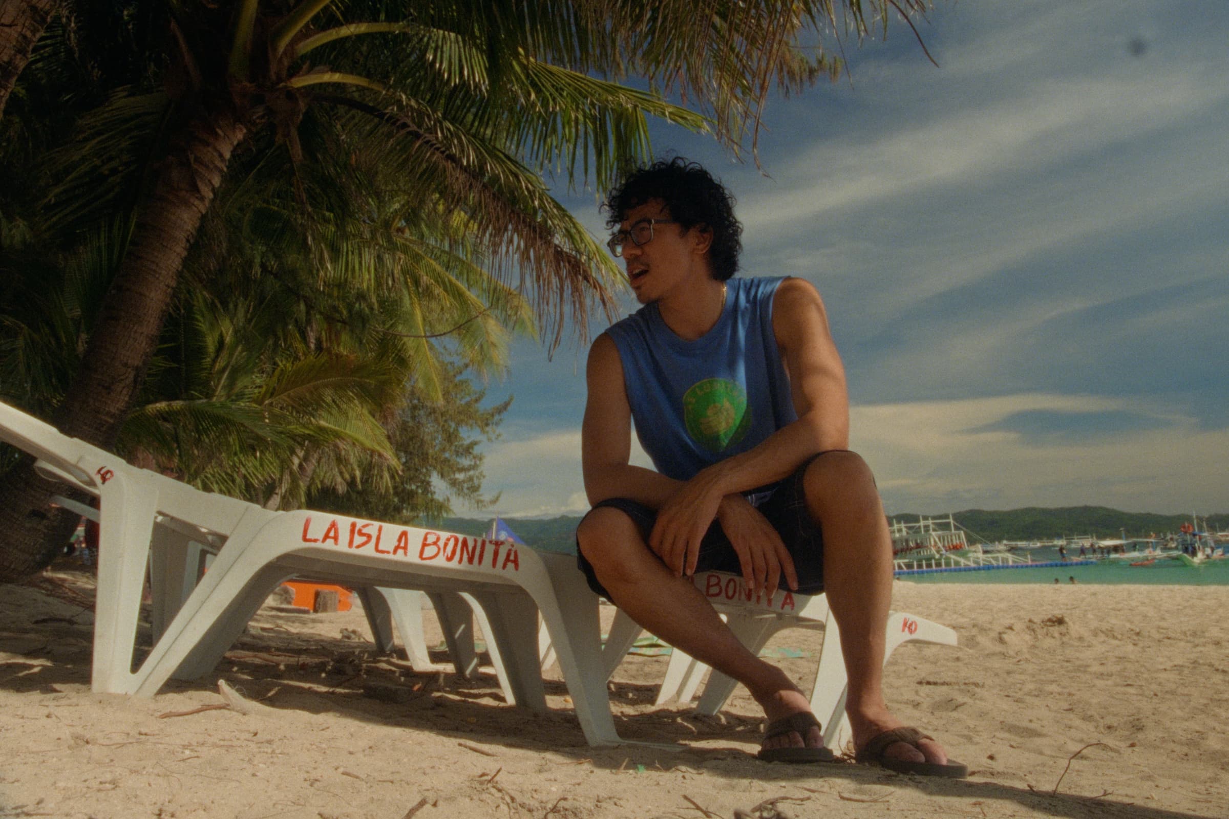 Andrew Amisola in White Sands in Boracay — Andrew sitting on a white plastic beach lounger stenciled LA ISLA BONITA in red, palm fronds overhead and the sea behind.