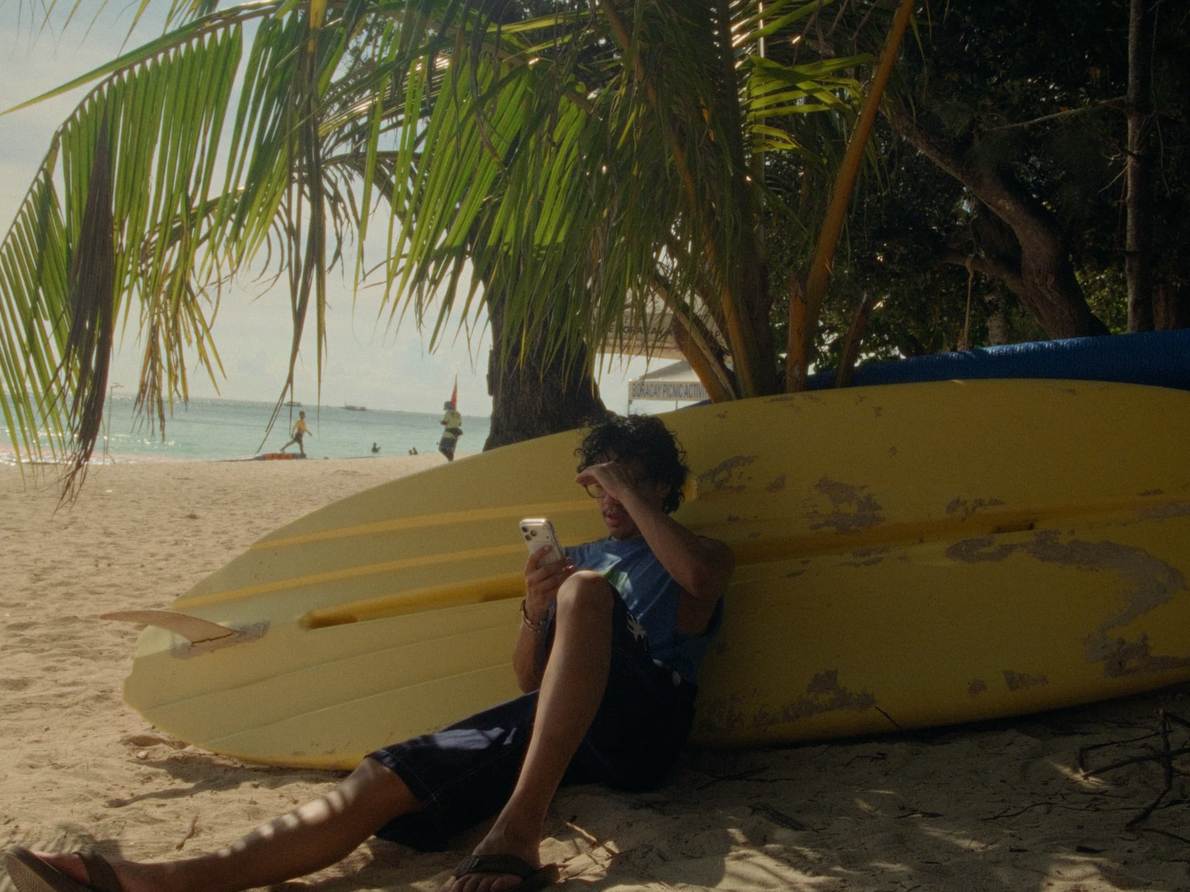 Andrew Amisola in White Sands in Boracay — Andrew sitting on the sand against the underside of a yellow surfboard, looking at his phone, palm fronds and the shoreline behind him.