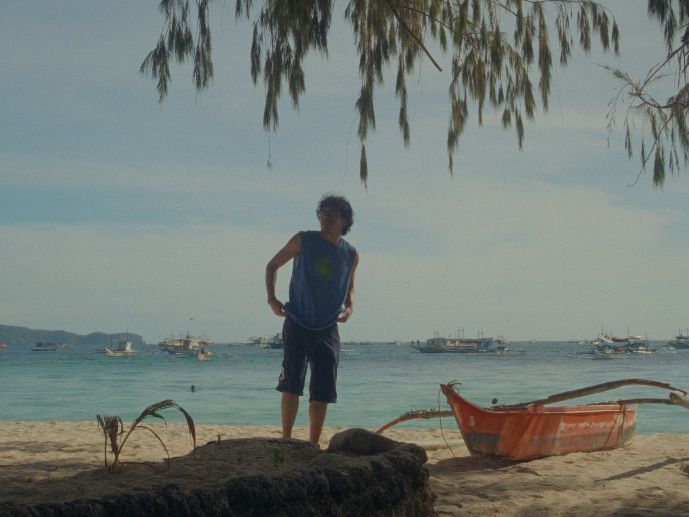 Andrew Amisola in White Sands in Boracay — Andrew standing on a low sandstone outcrop above the beach in a blue tank, an orange paraw and other anchored outriggers behind him.
