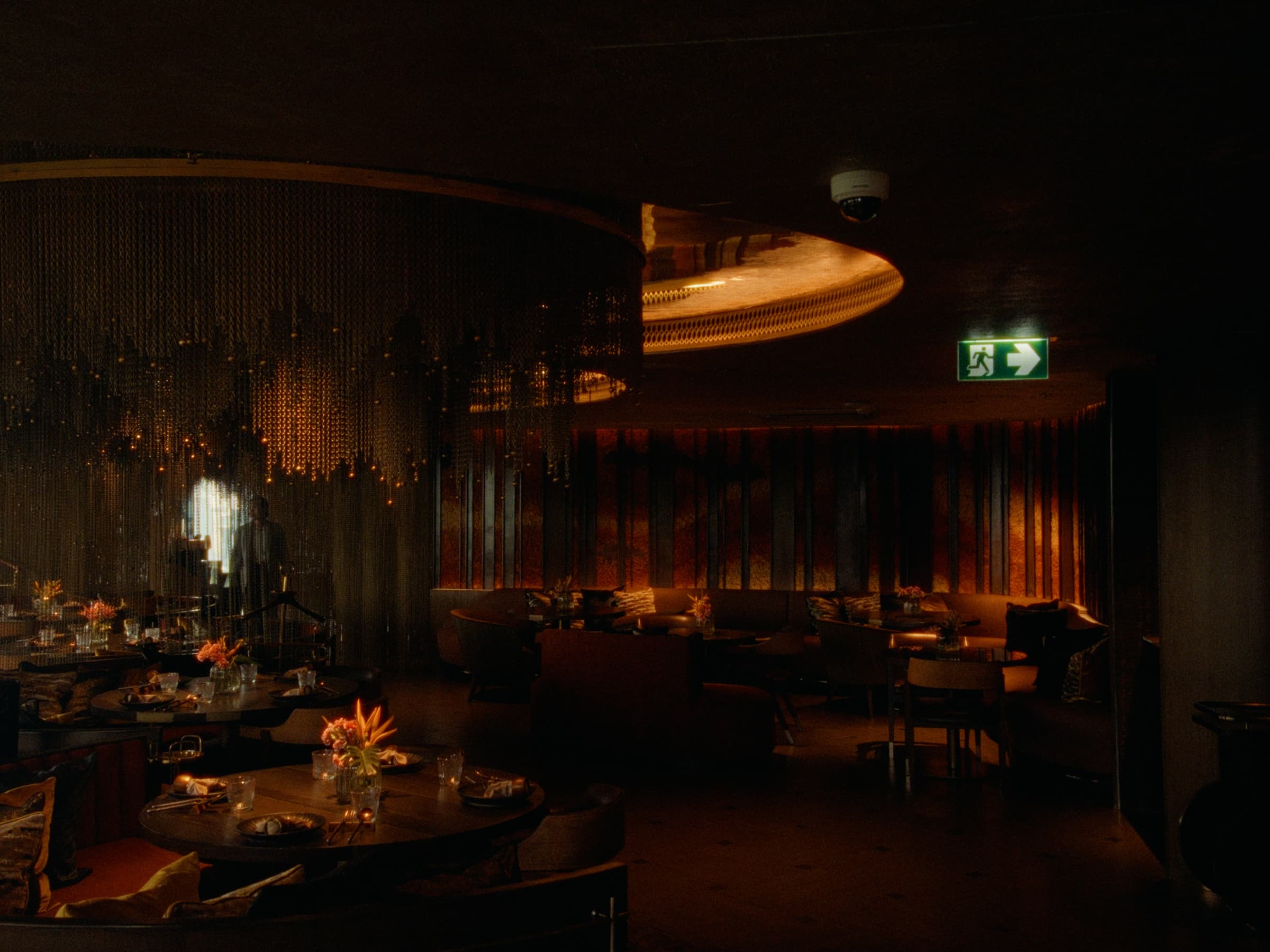 A dim Bangkok lounge interior lit by curtains of chain and a brass ceiling disc, with tables set for service and a green exit sign on the wall