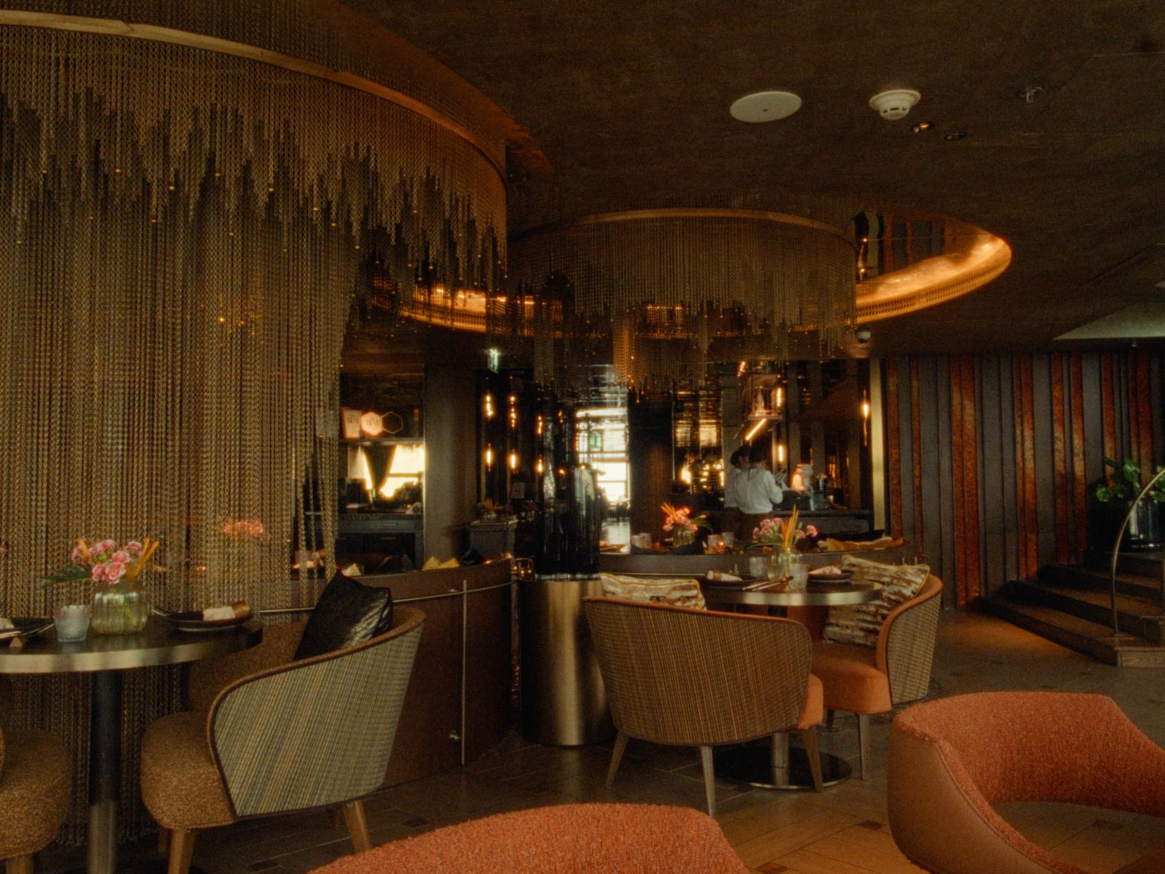 A second view of the same Bangkok restaurant — curtains of chain frame an open kitchen with staff and stacked wine displays beyond