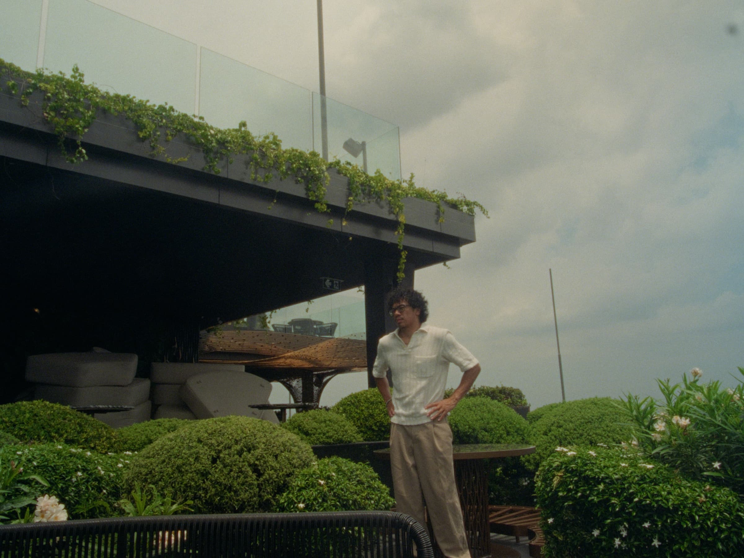 Andrew Amisola — Andrew on a Bangkok rooftop terrace surrounded by clipped boxwood spheres, ivy spilling over a cantilevered black canopy above