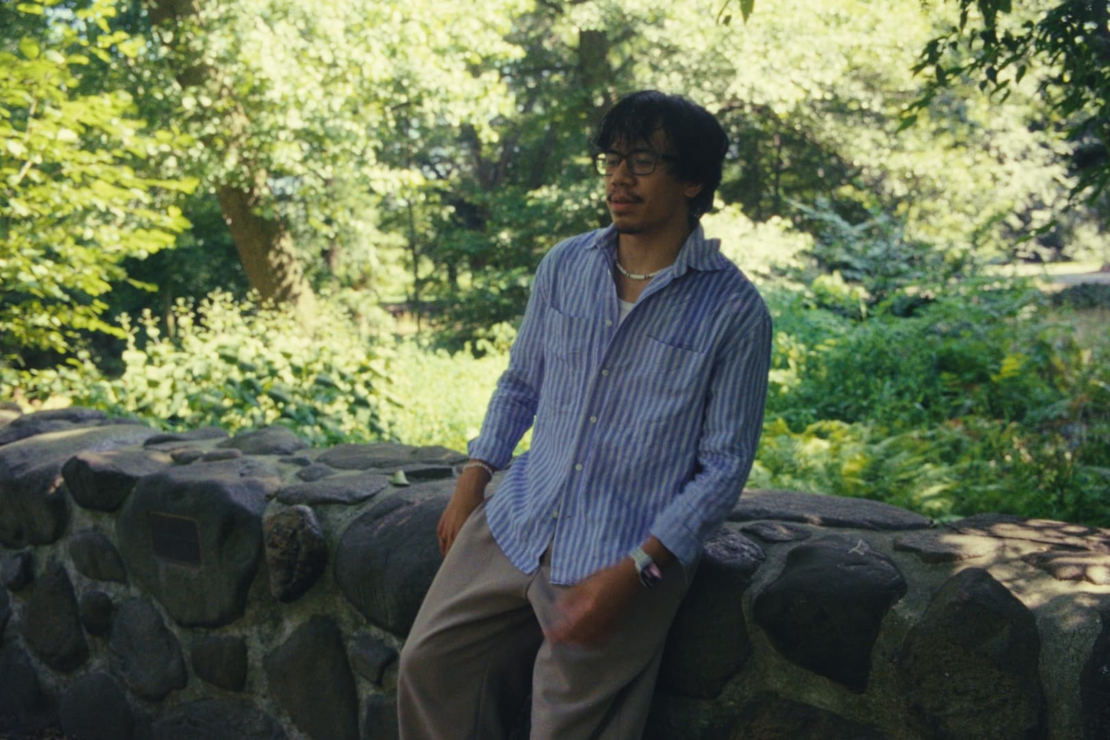 Andrew Amisola — Andrew seated on a low fieldstone wall in a blue-and-white striped shirt, surrounded by sunlit summer foliage at the Brooklyn Botanical Garden
