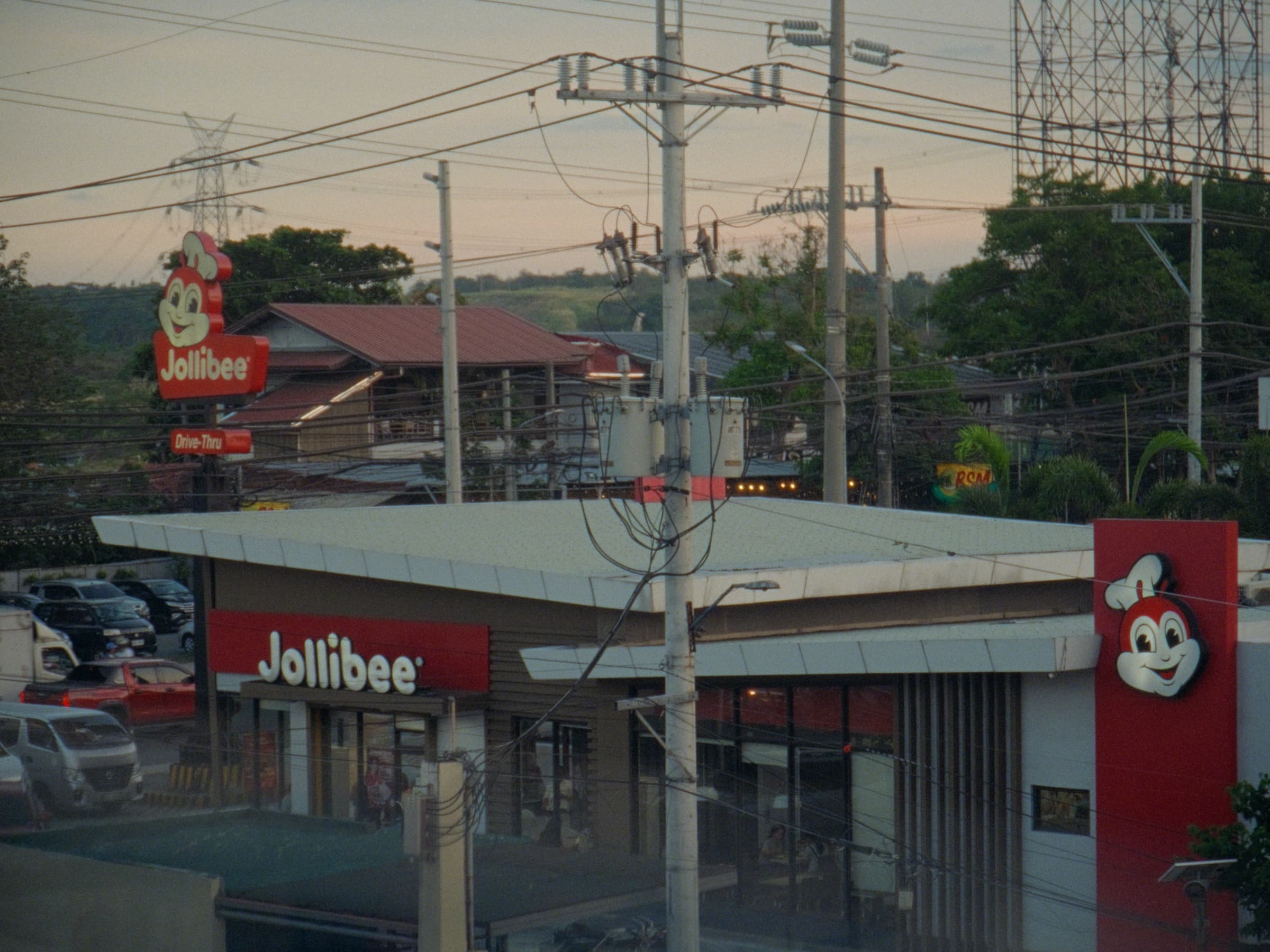 A Jollibee drive-thru storefront at golden hour with the mascot sign rising into a sky crisscrossed by power lines and pylons