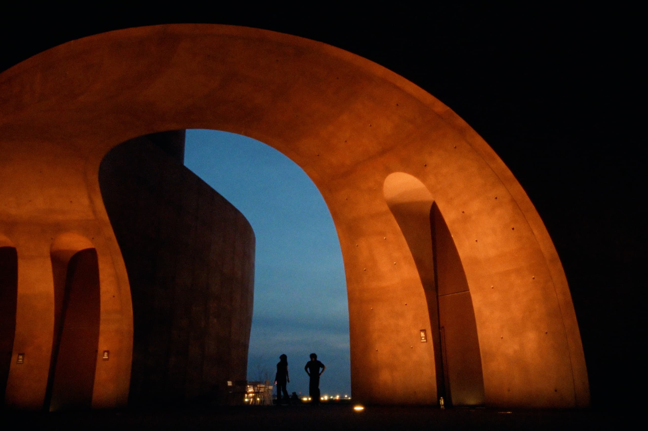 Two silhouettes standing beneath massive illuminated arches at dusk with twilight sky beyond