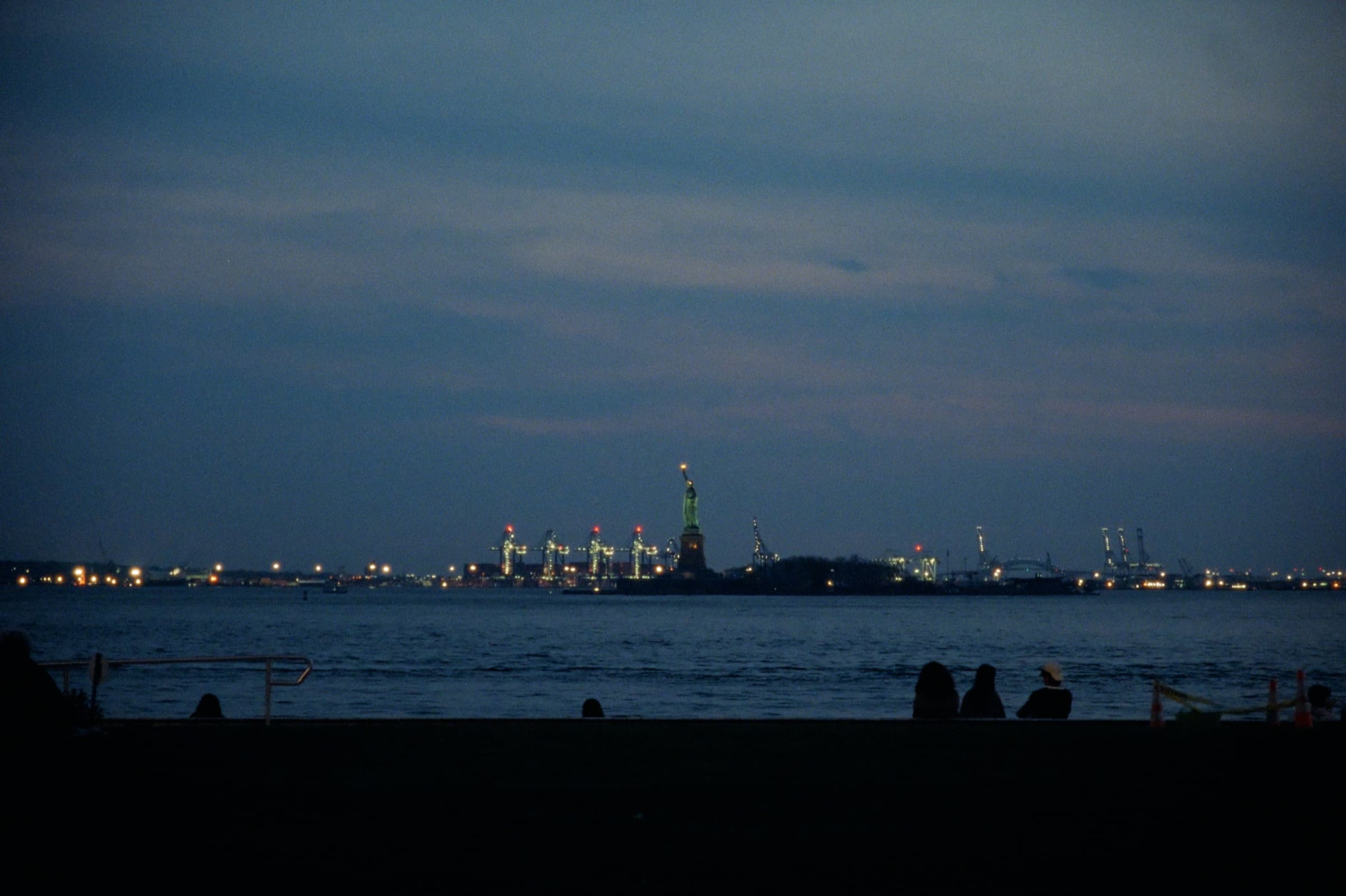 Andrew Amisola — Statue of Liberty across the harbor at dusk with port lights and silhouetted figures in the foreground