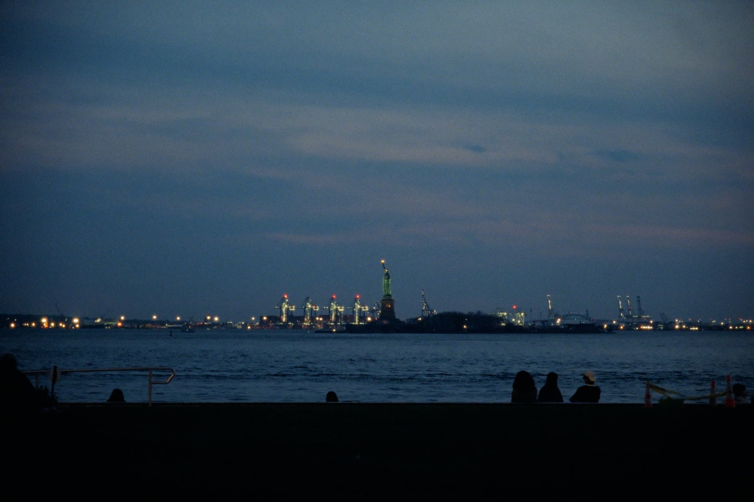 Statue of Liberty across the harbor at dusk with port lights and silhouetted figures in the foreground