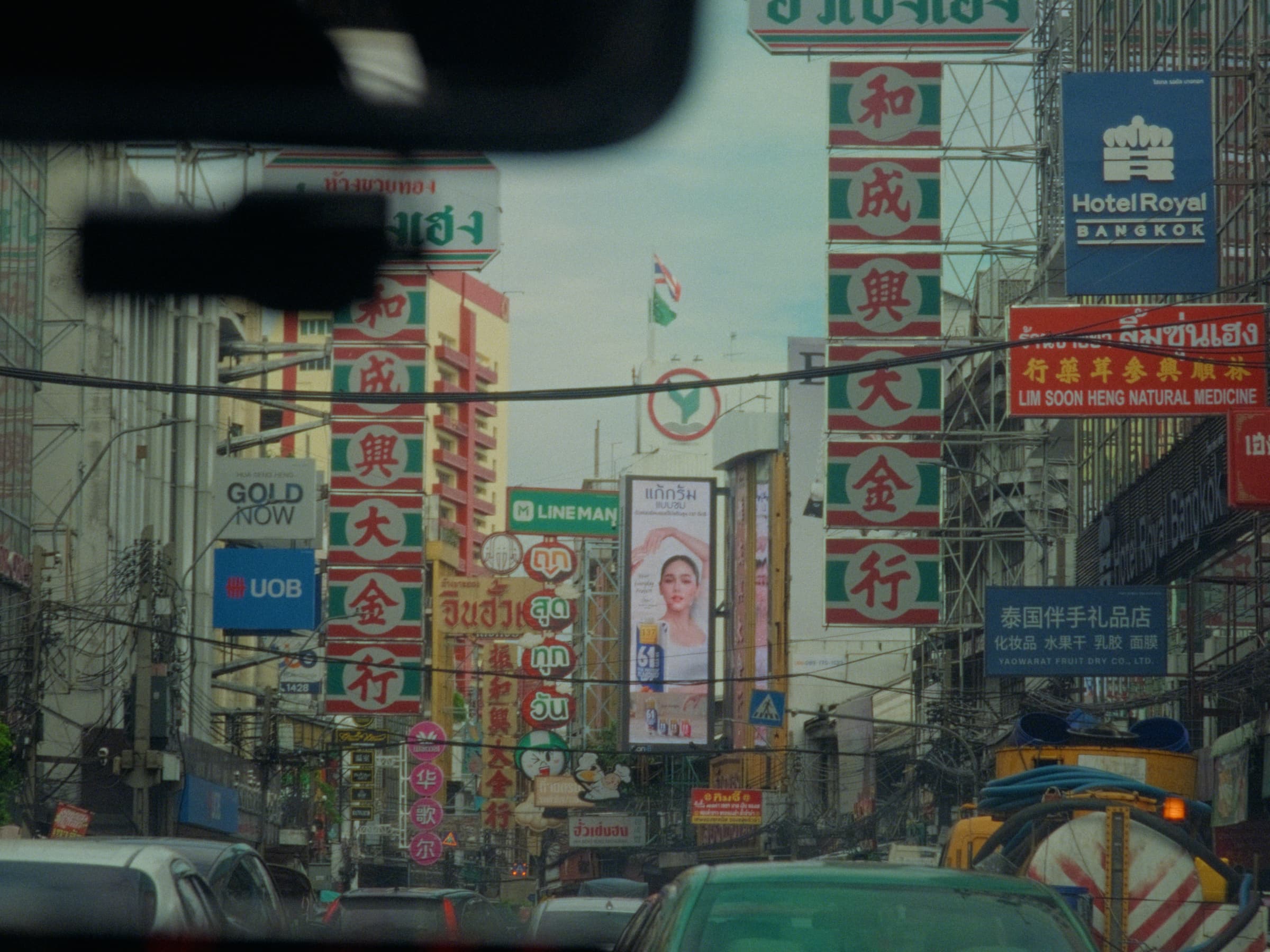 View through a car windshield down a Bangkok Chinatown street stacked with red-and-green Chinese signage and a Hotel Royal Bangkok placard