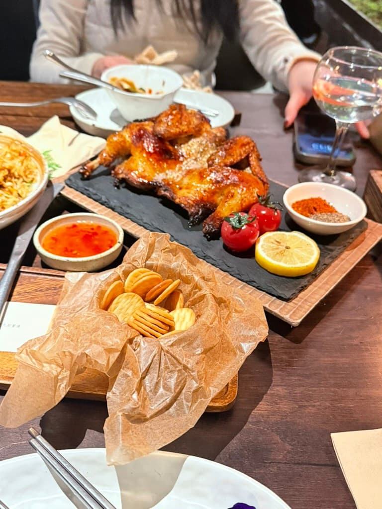 A whole roast chicken on a slate board with cherry tomatoes, a halved lemon, and chili seasoning, alongside crackers in parchment and a bowl of sweet chili sauce