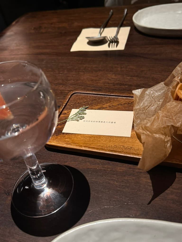 A wooden tray with a small card reading 世间所有的相遇都是久别重逢 beside a dried sprig, a glass of wine, and crackers wrapped in parchment