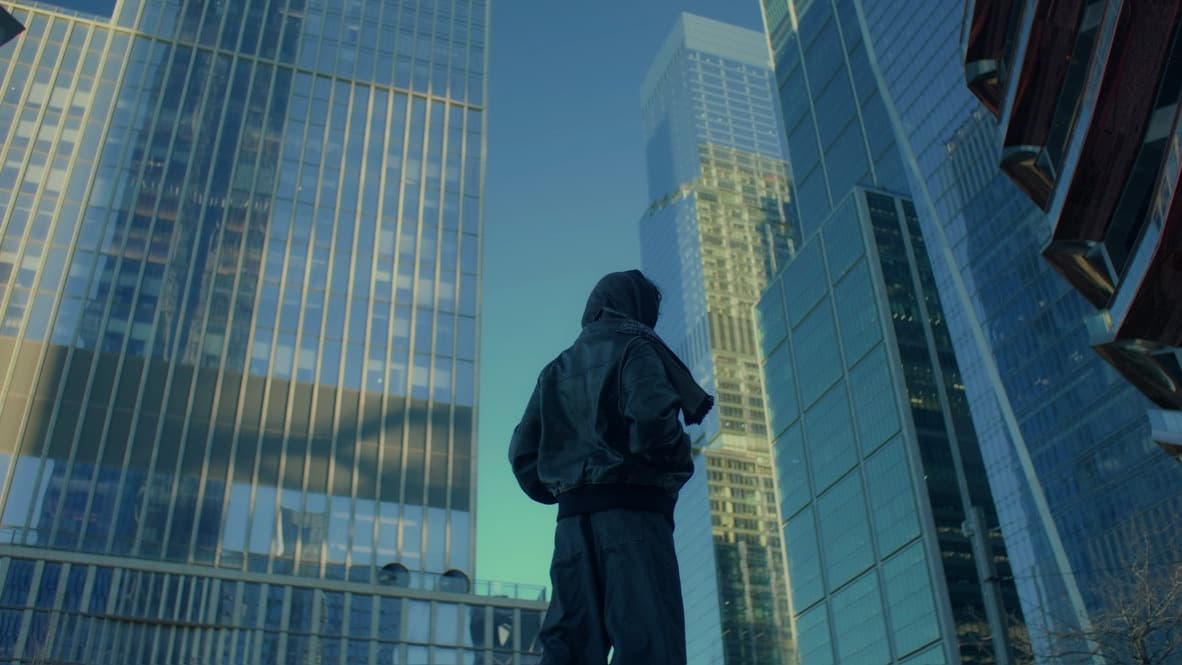 Andrew in a hood with his back to the camera, looking up at glass towers, a still from the Daylight Savings music video