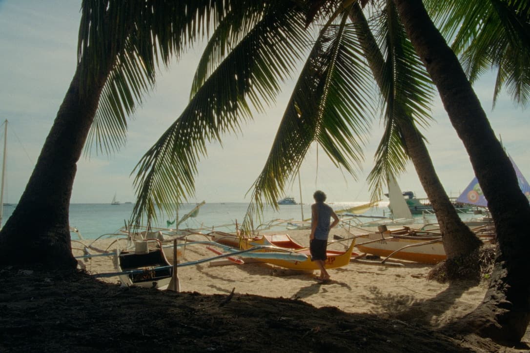 Andrew walking among outrigger boats on White Beach in Boracay, framed by leaning coconut palms