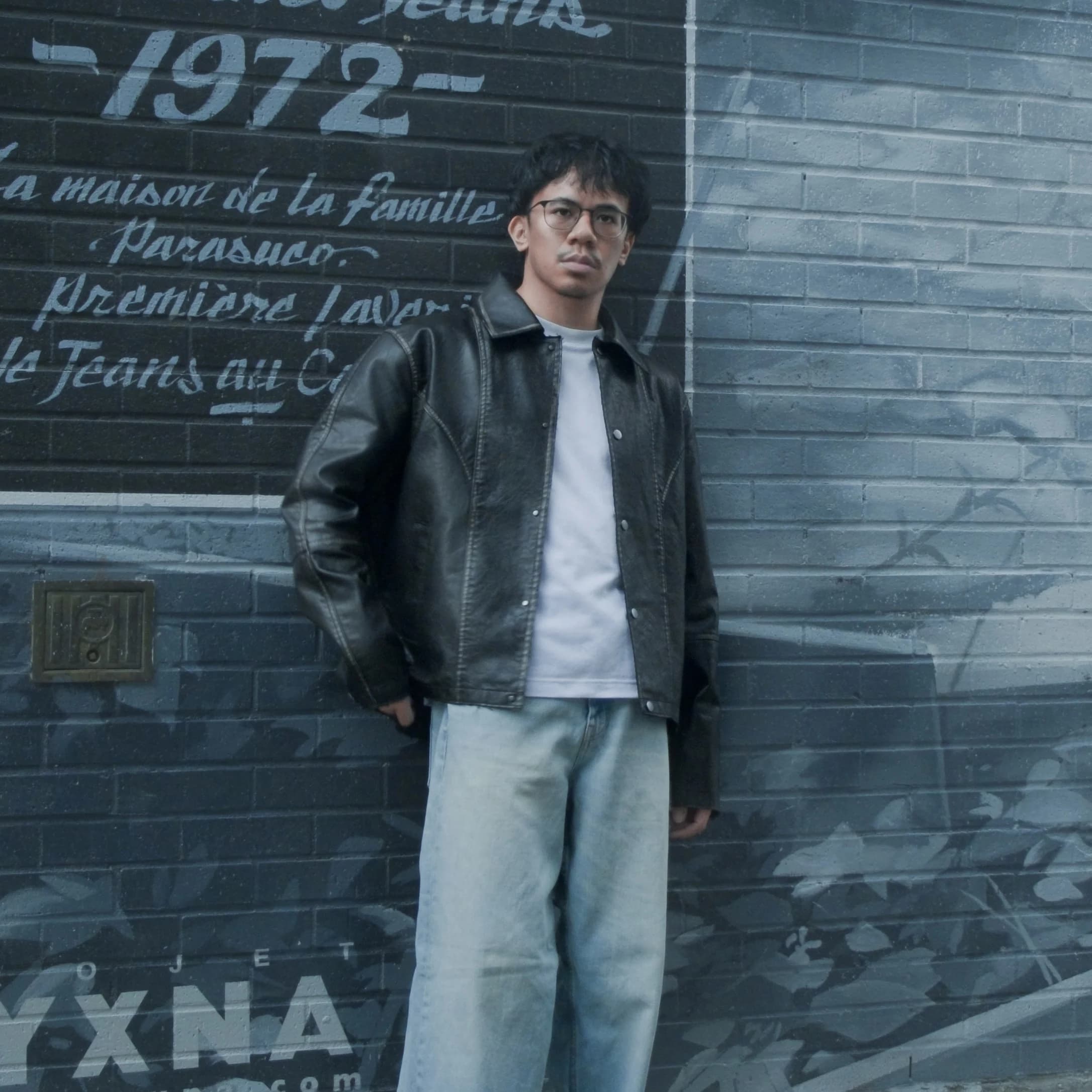 Andrew Amisola in a black leather jacket and jeans, standing against a blue painted mural that reads 'Parasuco Jeans, 1972'